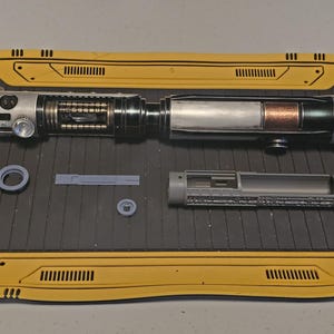 Op de afbeelding: Een gedemonteerde lichtzwaard prop met zilveren, zwarte en koperen componenten. Het handvat heeft gedetailleerde mechanische elementen en het bladgedeelte onthult interne structuren. Extra grijze onderdelen en gele panelen zijn ook aanwezig.