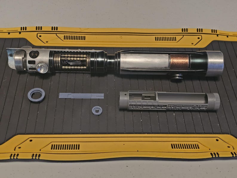 Op de afbeelding: Een gedemonteerde lichtzwaard prop met zilveren, zwarte en koperen componenten. Het handvat heeft gedetailleerde mechanische elementen en het bladgedeelte onthult interne structuren. Extra grijze onderdelen en gele panelen zijn ook aanwezig.