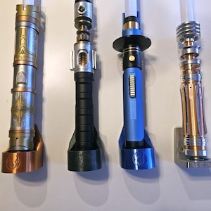 May include: Four lightsaber hilts mounted on a wall. The hilts are made of metal and have different colors and designs. The hilts are all in a vertical position and are evenly spaced apart. The hilts are all in focus and the background is blurred.
