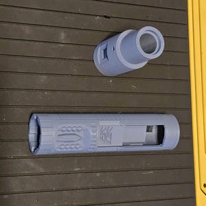 Puede incluir: Dos objetos cilíndricos azul claro impresos en 3D. Uno es un tubo hueco con patrones de superficie detallados y una abertura rectangular. El otro es una pieza cilíndrica más corta con una abertura circular. Ambos están sobre una superficie oscura y texturizada.