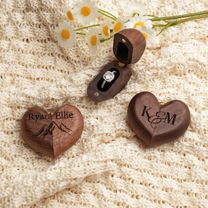 May include: A diamond engagement ring in a wooden ring box, with two wooden heart-shaped boxes. One heart is engraved with "Ryan Ellie" and mountain graphics. The other heart is engraved with the letters "K & M". The items are on a cream-colored knitted surface.
