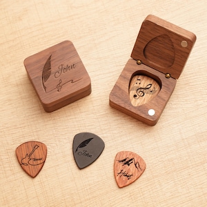 May include: Wooden guitar pick gift set. Includes a closed and open wooden box, each with a magnetic closure. The closed box has a feather and musical note design with the name "John". Three guitar picks with different designs are also included.