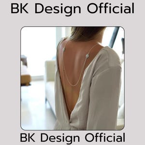 May include: A silver back chain necklace with two circular pendants. The necklace is worn by a person with a light gray top.