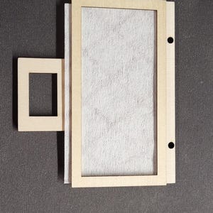 May include: A wooden picture frame with a rectangular opening, accompanied by a smaller square frame. The frame is made of light-colored wood and contains a textured, off-white material. Two small holes are drilled into the side of the frame.