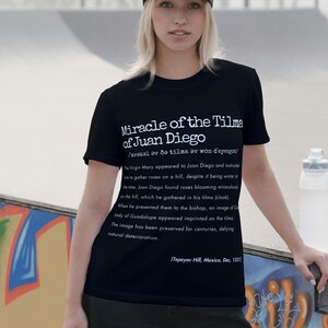 May include: Black t-shirt with white text describing the Miracle of the Tilma of Juan Diego. The text includes a phonetic pronunciation and a detailed account of the event, including the appearance of the Virgin Mary and the image of Our Lady of Guadalupe. The shirt's design is a historical and religious theme.