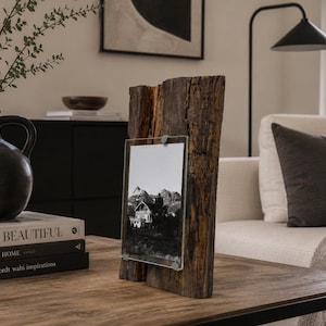May include: A vertical picture frame constructed from a piece of dark, weathered, textured wood with a rough bark edge. A clear glass pane is attached to the front, holding a black and white photograph of a mountain landscape with a small house.
