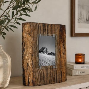 May include: A rectangular picture frame crafted from a thick, natural piece of dark brown wood with visible bark edges. A vertical glass pane is secured to the center of the wood by four metal clips, displaying a black and white photograph of a house.