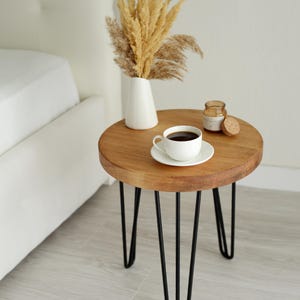 Peut inclure: Petite table d'appoint ronde en bois avec pieds épingles noirs. Une tasse et une soucoupe blanches, remplies de café noir, sont posées dessus. Un vase blanc avec des fleurs séchées et un petit pot avec un couvercle en liège sont également présents.