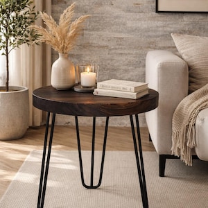 Puede incluir: Una mesa auxiliar redonda de color marrón oscuro con patas de horquilla negras. La mesa tiene libros, una vela encendida y un jarrón con flores secas. La mesa está sobre una alfombra de color neutro.