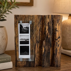 Puede incluir: Un marco de fotos rústico de madera con un acabado natural texturizado. El marco contiene tres fotografías de paisajes en blanco y negro, sujetas con clips metálicos. El marco se exhibe sobre una superficie de madera, con una lámpara y una planta en el fondo.