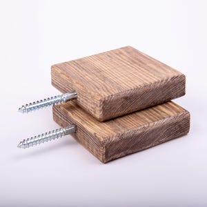 May include: Two stacked, rectangular wooden blocks with visible wood grain. Each block has a silver screw inserted. The wood has a natural, light brown color with darker brown streaks, suggesting a rustic or reclaimed aesthetic.