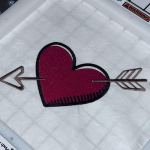 Puede incluir: Un aro de bordado blanco con un corazón rojo y una flecha marrón con rayas blancas. La flecha atraviesa el corazón. El texto "Strong Magnets" y "AWARNING" es visible en el aro.