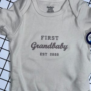 以下が含まれることがあります： 前面にピンクで「FIRST Grandbaby EST 2025」と刺繍された白いベビーロンパースです。