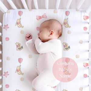 Puede incluir: Una sábana blanca con un estampado de Winnie the Pooh, globos rosas, lazos, estrellas y lunares. Un bebé con un body blanco duerme sobre la sábana. El texto "Custom Baby's Name" está en un círculo rosa.