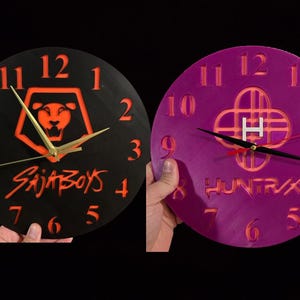 May include: Two round wall clocks. One is black with orange numerals and a lion logo, the text "SAJABOYS", and gold hands. The other is purple with orange numerals, a logo with the letter "H", and black hands.