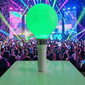 May include: A glowing green LED light stick with a white handle, resting on a white surface. The background shows a concert with a crowd of people holding similar lights, illuminated by colorful stage lights and confetti.
