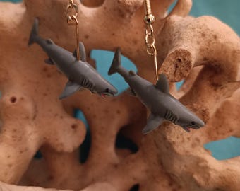 Pode incluir: Brincos de tubarão. Cada brinco apresenta um pequeno pingente de tubarão cinza suspenso por um gancho dourado. Os tubarões têm barbatanas detalhadas e um detalhe vermelho para a boca. Os brincos são exibidos em um fundo bege texturizado.
