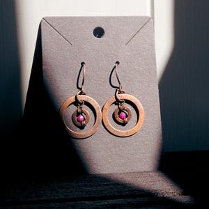 May include: A pair of copper-toned dangle earrings featuring a circular metal frame with a smaller inner ring. Each earring is centred with a small, round, vibrant pink bead secured by thin wire wrapping, suspended from a curved metal hook.