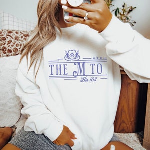 May include: White sweatshirt with a purple graphic design that says "The M to His 103". The graphic includes a heart with a star and wings.