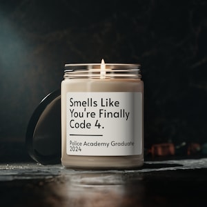 Peut inclure: Une bougie brune avec une étiquette blanche qui indique "Smells Like You're Finally Code 4. Police Academy Graduate 2024". La bougie est allumée et la flamme est visible.