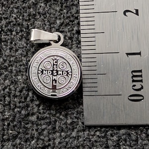 May include: A silver pendant with a circular design and the text "C S N D S M D P B" and other text. The pendant is next to a ruler showing centimetres.
