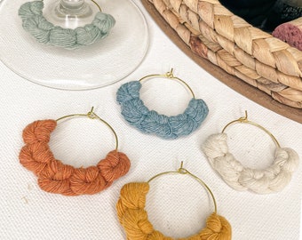 May include: Four handmade macrame hoop earrings in different colors: sage green, blue, orange, and yellow. The hoops are made with a braided cord and have a gold metal hoop.