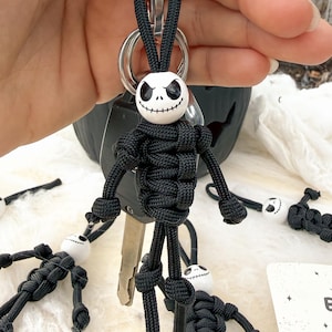 May include: A keychain featuring a figure made of black paracord with a white spherical bead head. The head has a painted black face with hollow eyes and a stitched mouth. The body is constructed from knotted cord, with limbs ending in knots.