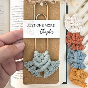 以下が含まれることがあります： 「Just One More Chapter」という文字と青いマクラメのタッセルが付いた金のブックマーク。ブックマークは、白い縁取りのある茶色の背景の上に置かれています。