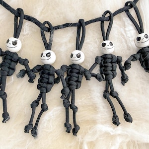 May include: A garland featuring five small figures made from black paracord knots with white spherical heads. Each head is painted with a black skeletal face design. The figures are suspended from a black cord, creating a series of hanging ornaments.