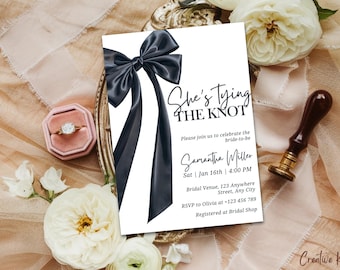 May include: A white wedding invitation with a black satin bow and the text "She's Tying The Knot" in black script. The invitation includes the bride-to-be's name, the date and time of the wedding, the venue, and RSVP information.