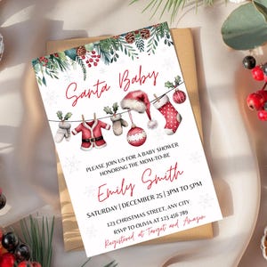 Puede incluir: Una tarjeta blanca con una ilustración de acuarela roja y verde del gorro, los guantes y una media de Papá Noel colgados de una cuerda para tender la ropa. El texto "Santa Baby" está escrito en cursiva roja. La tarjeta es una invitación a una fiesta de bienvenida al bebé para Emily Smith, con la fecha y la hora de la fiesta.