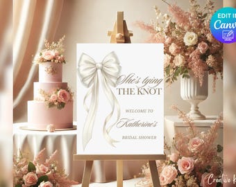 May include: White bridal shower welcome sign with a large white satin bow and the text "She's tying the knot" and "Welcome to Katherine's Bridal Shower".