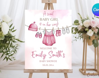 Op de afbeelding: Een roze welkomstbord voor een babyshower met een aquarelontwerp van babykleding die aan een waslijn hangt. De tekst luidt "A sweet BABY GIRL is on her way!" en "Welcome to Emily Smith's Baby Shower 16.01.20xx".
