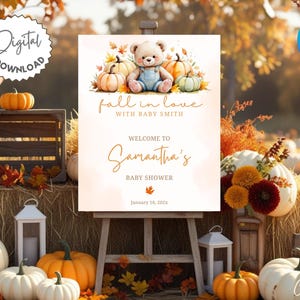 Pode incluir: Uma placa de chá de bebê com um ursinho de pelúcia e abóboras. A placa diz "fall in love with Baby Smith" e "Welcome to Samantha's Baby Shower". O fundo apresenta abóboras, fardos de feno e folhagem de outono.