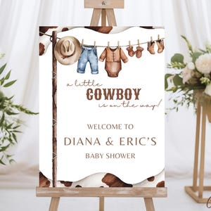 May include: A white sign with brown text announcing a baby shower for Diana & Eric. The sign says "a little COWBOY is on the way!" and features a line of baby clothes hanging on a clothesline, including a cowboy hat, jeans, a onesie, and booties.