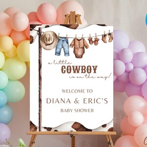 May include: A white sign with brown accents and a cow print background. The sign says "a little COWBOY is on the way!" and "WELCOME TO DIANA & ERIC'S BABY SHOWER".