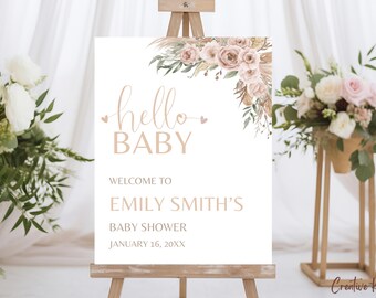 May include: A baby shower welcome sign on a wooden easel. The sign reads "hello BABY" in elegant script, with the name "EMILY SMITH'S" and the date "JANUARY 16, 20XX". Floral accents in soft pink and beige adorn the top right corner.