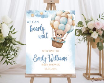 May include: A baby shower sign with a teddy bear in a hot air balloon, the text reads "We can bearly wait" and "Welcome to Emily Williams Baby Shower". The sign is on a wooden easel, with floral arrangements on either side.
