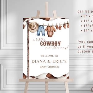 May include: A white sign with a brown cowhide pattern border. The sign says "a little COWBOY is on the way!" and "WELCOME TO DIANA & ERIC'S BABY SHOWER". There are illustrations of baby clothes hanging on a clothesline. The sign also includes information about printing sizes: 8" x 10", 11" x 13.75", 16" x 20", and 24" x 30".