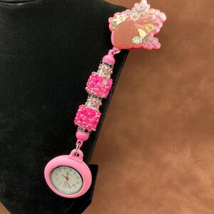 May include: Pink beaded retractable badge reel with a pink pocket watch and a heart-shaped charm with floral details. The charm has a glittery finish.