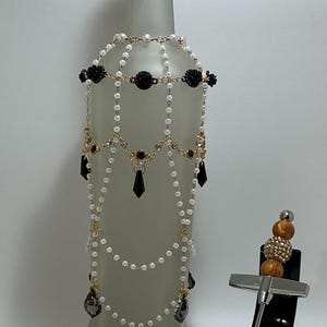 May include: A decorative wine bottle with a green top and a beaded embellishment. The embellishment features white pearls, black floral accents, and dangling black teardrop-shaped beads. A wine bottle stopper and corkscrew are also displayed.