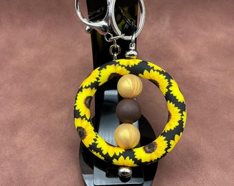 May include: A sunflower-themed keychain with a black and yellow floral pattern. The keychain features a large, round silicone bead with a sunflower design, and three smaller wooden beads in gold and brown. The keychain has a silver metal clasp.