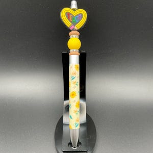 May include: A decorative pen with a yellow heart-shaped topper that reads "TEACH" in colorful letters. The pen features a floral pattern on a cream background and is accented with yellow and brown beads.
