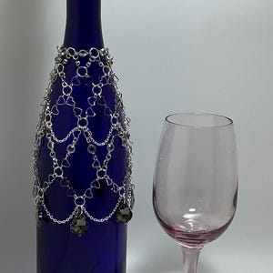 May include: A dark blue wine bottle with a decorative silver chain design and a wine glass. The chain has heart-shaped and round links, with dark gray crystal pendants. The wine glass is clear with a pink stem and bowl.