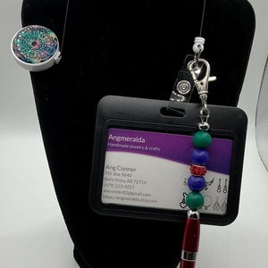May include: A black and white ID badge holder with a purple and white business card inside. The business card has the text "Angmeralda Handmade jewelry & crafts" and contact information for Ang Conner. A silver retractable badge reel with a colorful beaded charm is attached to the holder.