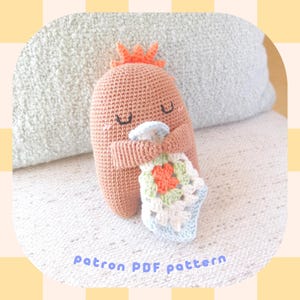 Puede incluir: Un juguete amigurumi de crochet con forma de monstruo marrón que sostiene una manta de crochet multicolor. El juguete tiene una pequeña corona naranja en la cabeza y los ojos cerrados. El juguete está sentado sobre una superficie texturizada de color azul claro y blanco. El texto "Patron PDF pattern" está escrito en morado debajo del juguete.
