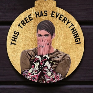 May include: A gold-coloured Christmas bauble featuring a portrait of a person covering their mouth with their hands. The bauble has the text "THIS TREE HAS EVERYTHING!" in black. It hangs against a dark wood background with Christmas tree branches.