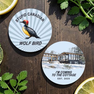 May include: Two round coasters on a wooden surface. One coaster features a cartoon bird with the text "STUPID CANADIAN WOLF BIRD." The other coaster has a picture of a modern house with the text "I'M COMING TO THE COTTAGE EST. 2025."