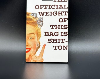 May include: A rectangular luggage tag with a vintage-style illustration of a woman pointing. The text reads "THE OFFICIAL WEIGHT OF THIS BAG IS SHIT-TON". The tag has a hole at the top for attachment.