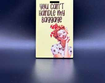 May include: A yellow luggage tag with a retro-style illustration of a woman with red hair and a red and white polka dot dress. The text on the tag reads "You can't handle my baggage."